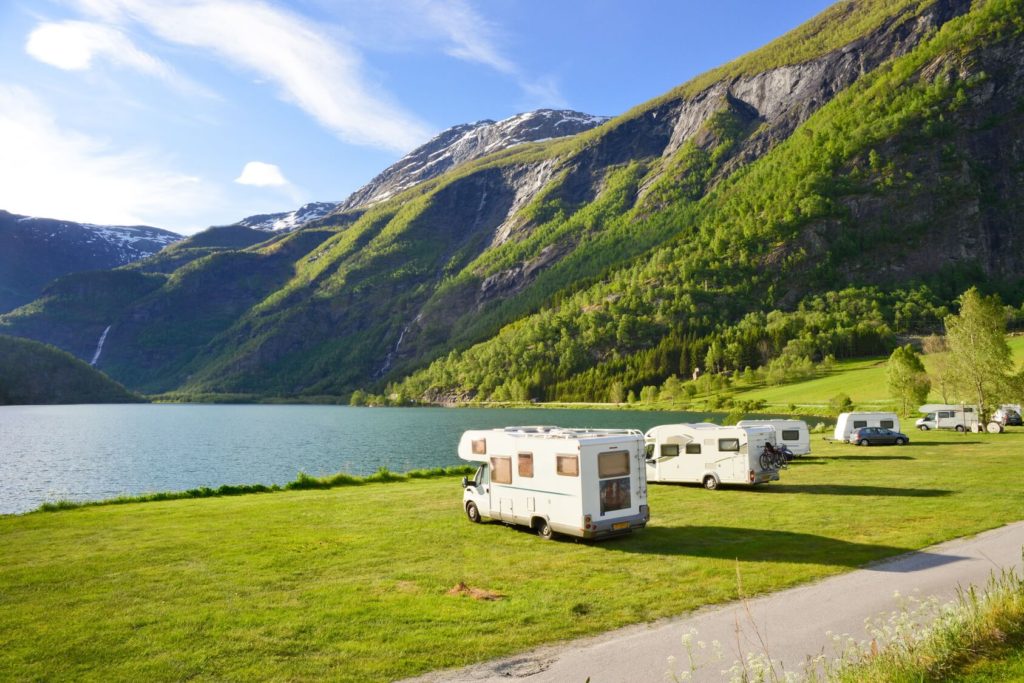 Mehrere Wohnmobile stehen auf einer grünen Wiese in der Sonne an einem See und Bergen.
