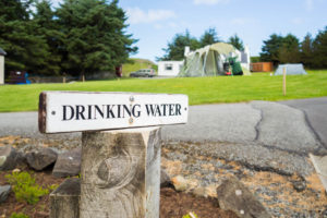 Ein weißes Schild mit der Aufschrift »Drinking Water« vor einem Campingplatz.