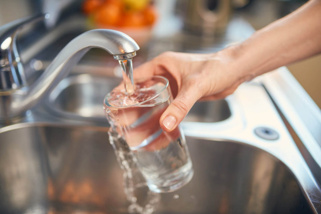 Eine Hand hält ein Glas Wasser unter den laufenden Wasserhahn eines Spülbeckens.